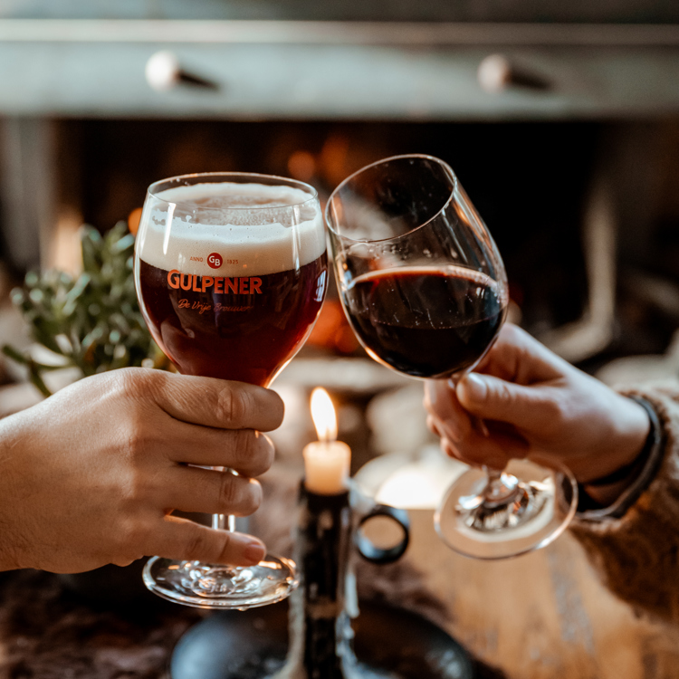 Bierglas en wijnglas proosten met aangestoken kaars in de achtergrond