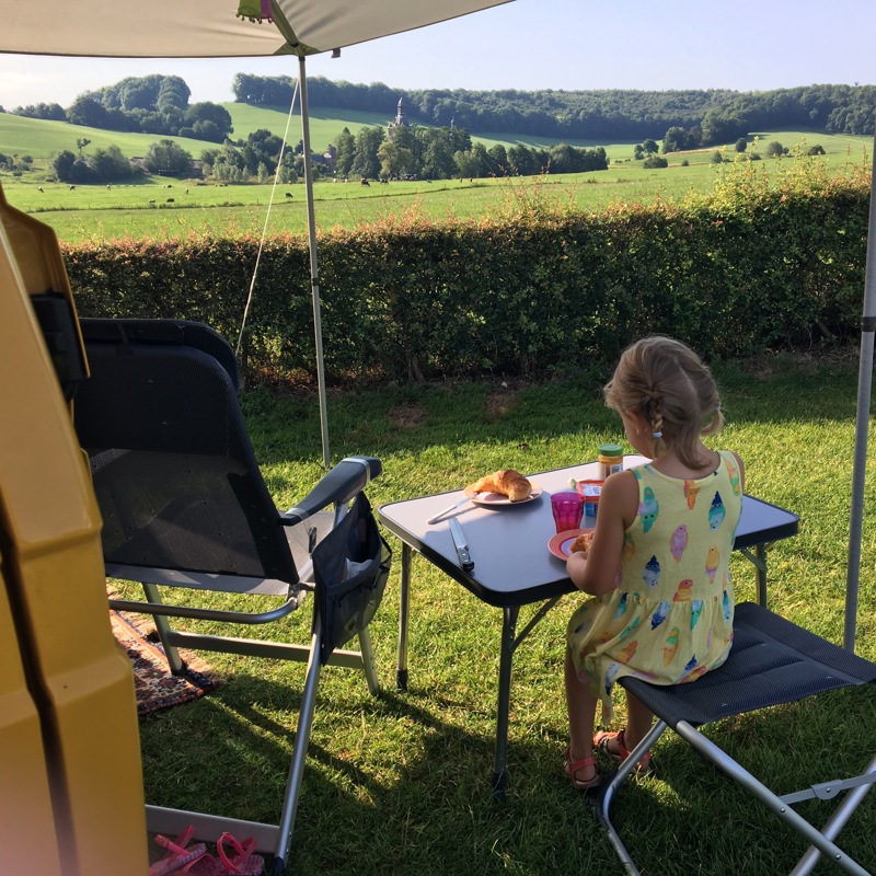 Meisje zit aan een kleine tafel met een croissantje op de camping. 