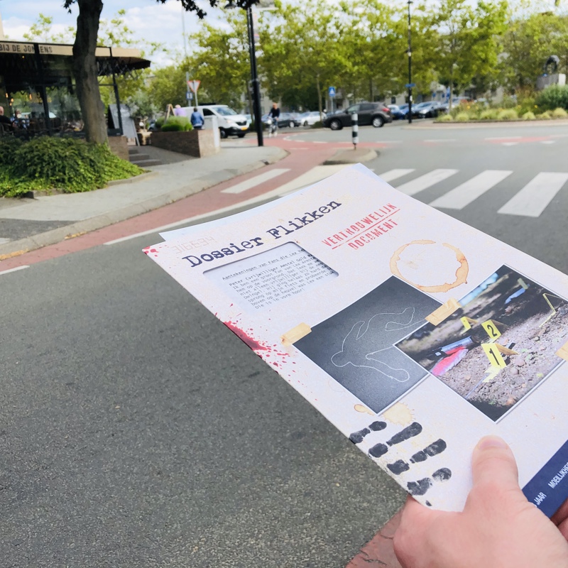 Straatbeeld met daarbij handen die eenn map met daarop 'Dossier Flikken' vasthouden