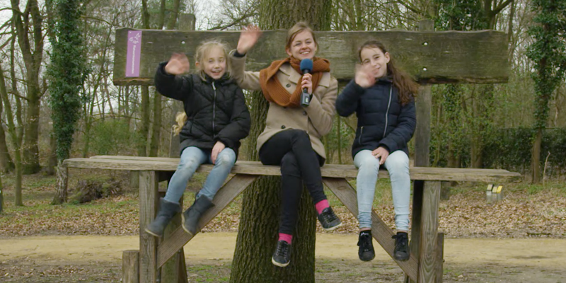 Presentatrice met 2 jonge meisjes op hoge houten bank tegen boomstam