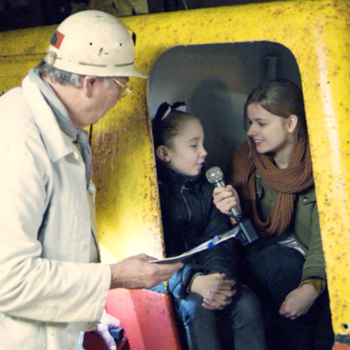 Koempel kijkende naar presentatrice die meisje interviewt in geel treintje in steenkolenmijn