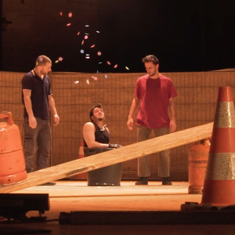 Theaterstuk met 3 mannen met op het podium een wip gemaakt van 2 oranje gasflessen en een houten plank