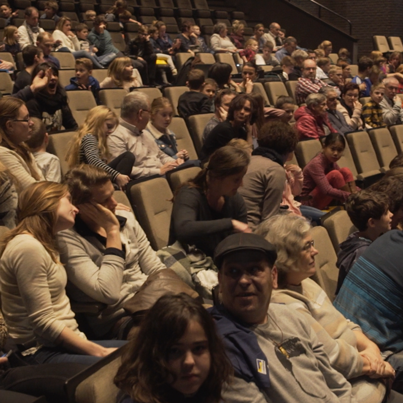 Publiek kijkende om zich heen in een theaterzaal waarbij enkel stoelen nog leeg zijn