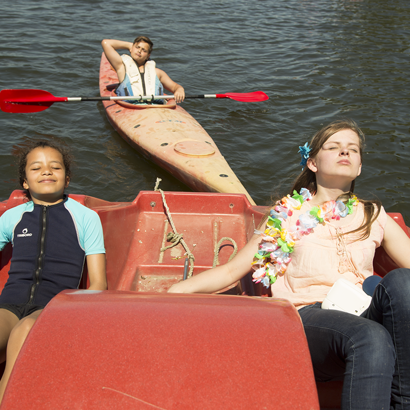 Jonge vrouw met hawaiketting geniet met meisje van zon in rode waterfiets. Achter hen doet jongen in kano hetzelfde