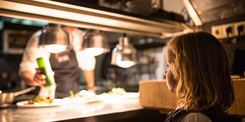 Kind staat in de keuken van een restaurant en bekijkt hoe de koks het eten bereiden