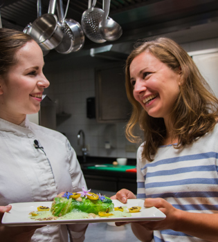 Een dame en de kokkin lachen naar elkaar en houden beide een bord met exclusief gerecht vast