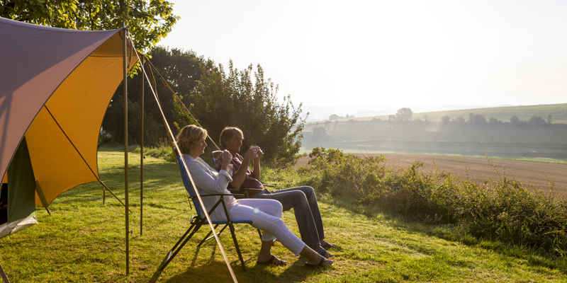 Echtpaar zit op twee campingstoelen voor de tent met een kopje koffie te genieten van de zon en het uitzicht. 