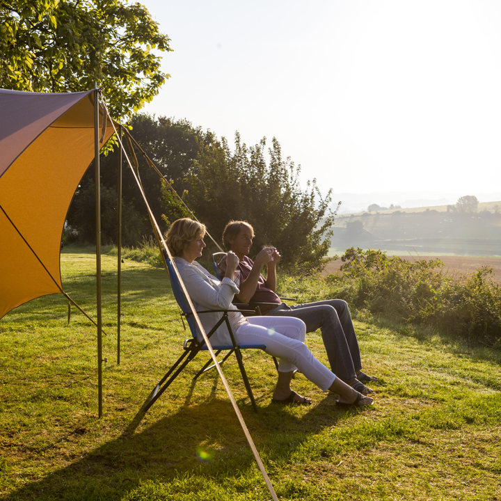 Echtpaar zit op twee campingstoelen voor de tent met een kopje koffie te genieten van de zon en het uitzicht. 