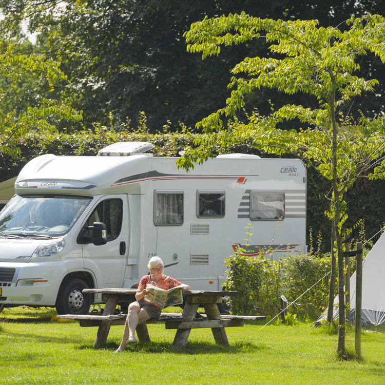 Vrouw zit krant te lezen op picknickbank met achter haar een grote camper. 