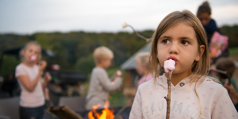 Meisje blaast op marshmallow op stokje, met daarachter andere kinderen en een vuurkorf. 