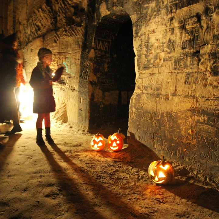 Mensen en kind staan voor gang in donkere grot met op de grond verlichte Halloween pompoenen