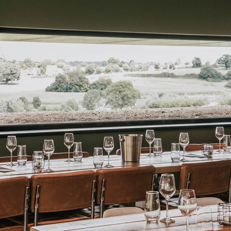 Cognac leren stoelen aan lange houten tafels met daarop lege water- en wijnglazen met uitzicht over het Heuvelland