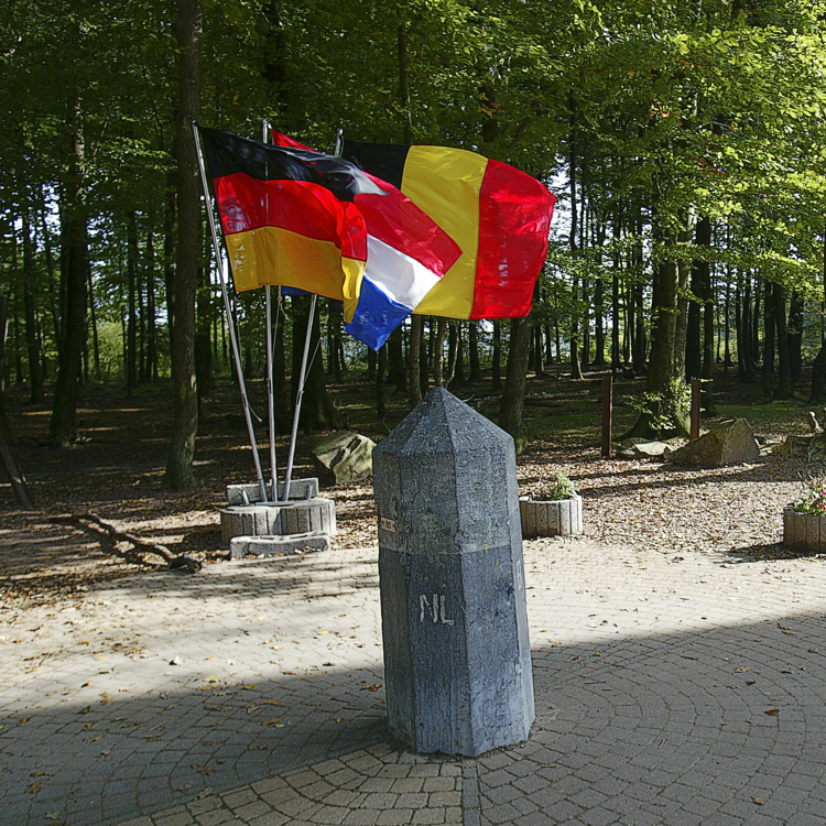 Natuurstenen grenspaal met daarachter de wapperende vlaggen van Duitsland, Nederland en België