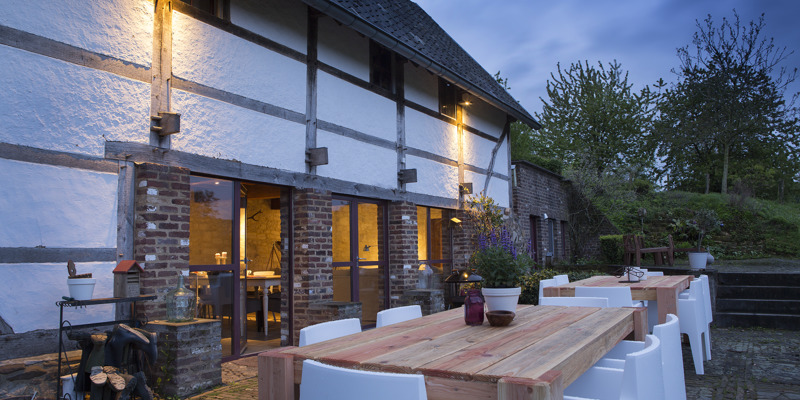 Groot terras met witte stoelen en houten tafels bij schemering.