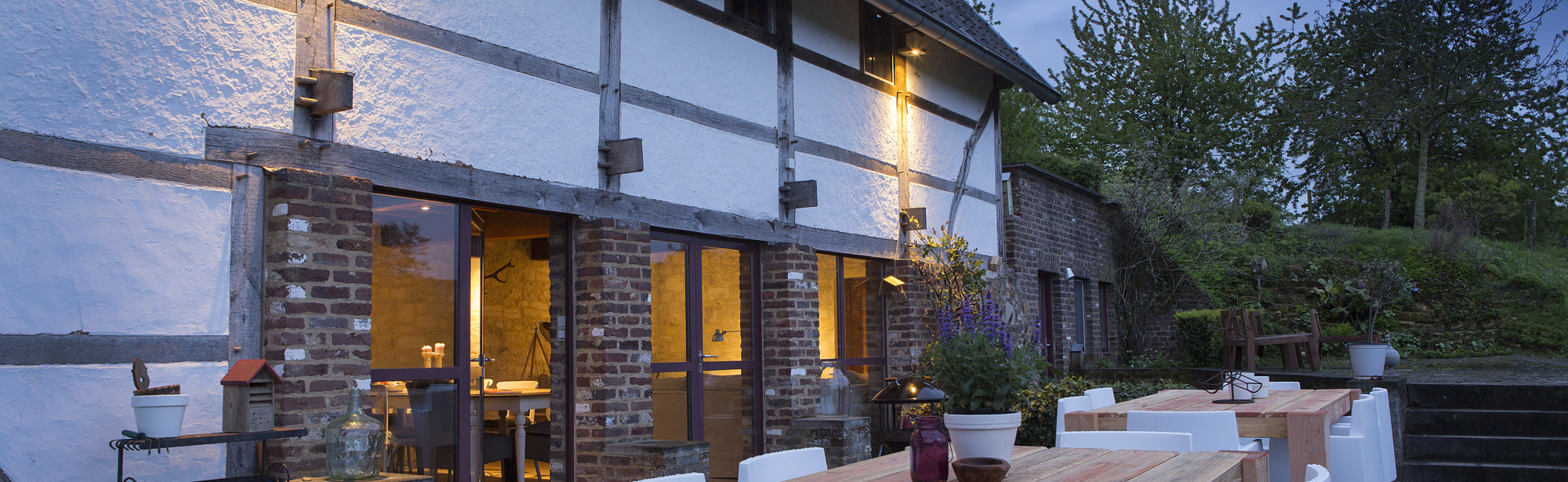 Groot terras met witte stoelen en houten tafels bij schemering.