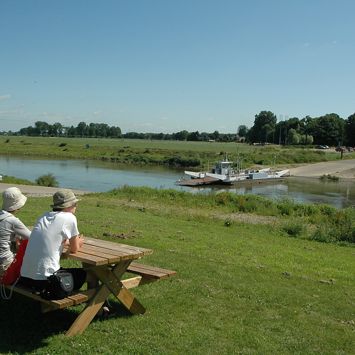 Twee personen aan een picknicktafel kijken uit over de maas naar het veerpontje