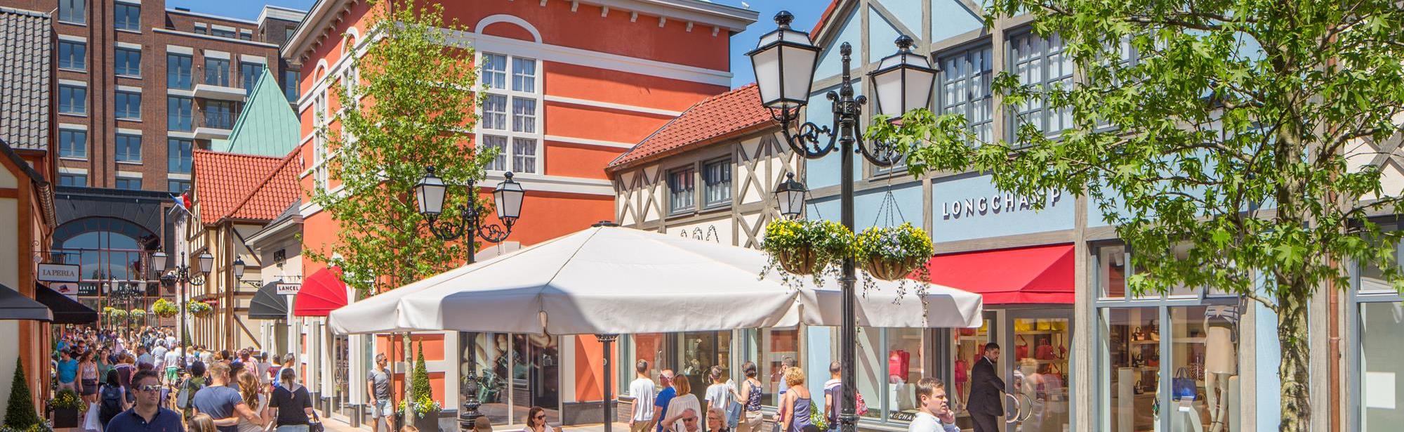 Mensen lopen met winkeltasjes door Maasmechelen Village of rusten uit op een bankje tijdens een zonnige dag