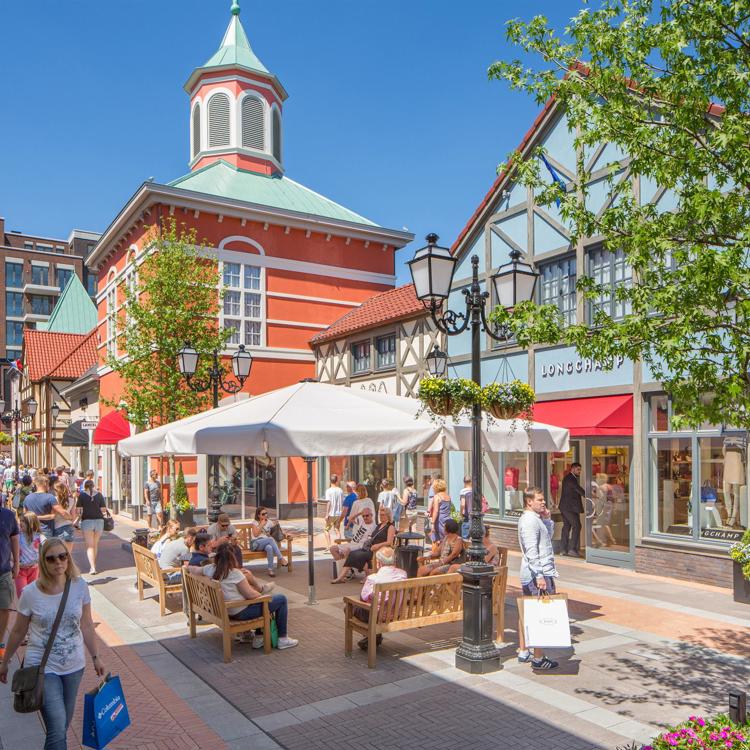 Mensen lopen met winkeltasjes door Maasmechelen Village of rusten uit op een bankje tijdens een zonnige dag