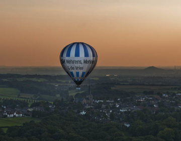 Blauw witte luchtballon die over Zuid-Limburg vliegt met een oranje lucht op de achtergrond