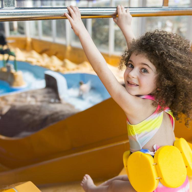 Kind met bruine krullen en gele drijfkurken gaat van de glijbaan af in het zwembad