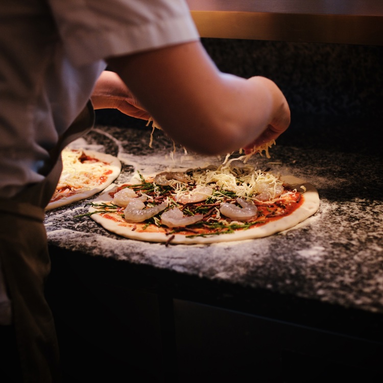 Een kok voegt de laatste kaas toe aan een pizza met garnalen, tomatensaus en groenten