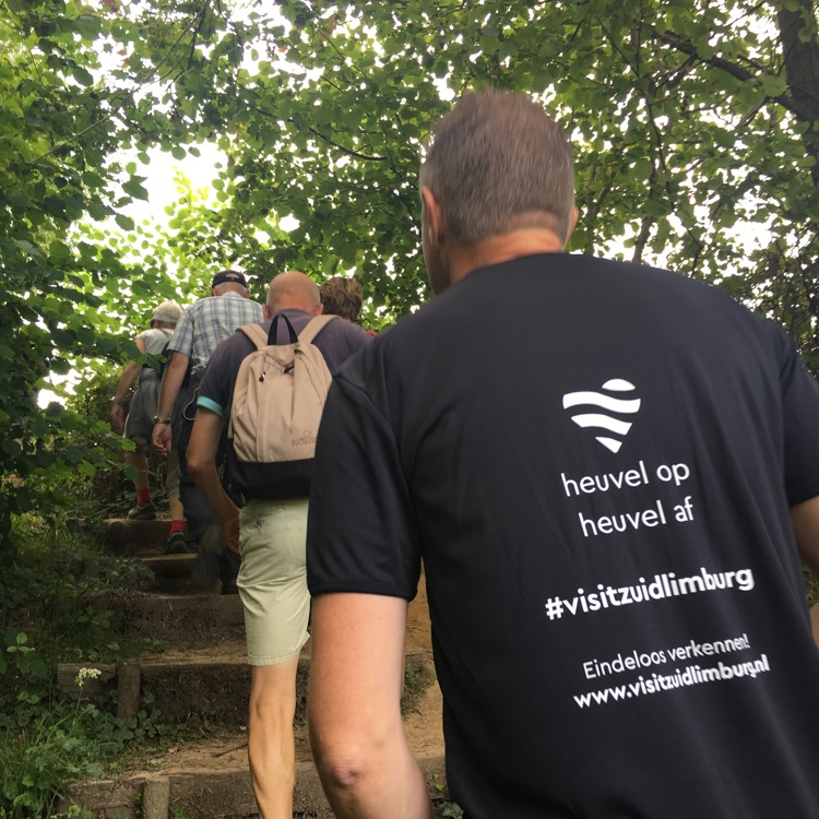 Wandelaars op trappen in een bus met man met bedrukt t-shirt met 'heuvel op, heuvel af. #visitzuidlimburg'