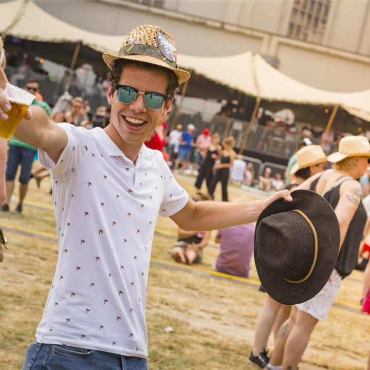 Jongeman met hoedje op zijn hoofd en hoed in zijn ene hand en een biertje in zijn andere op een festivalterrein