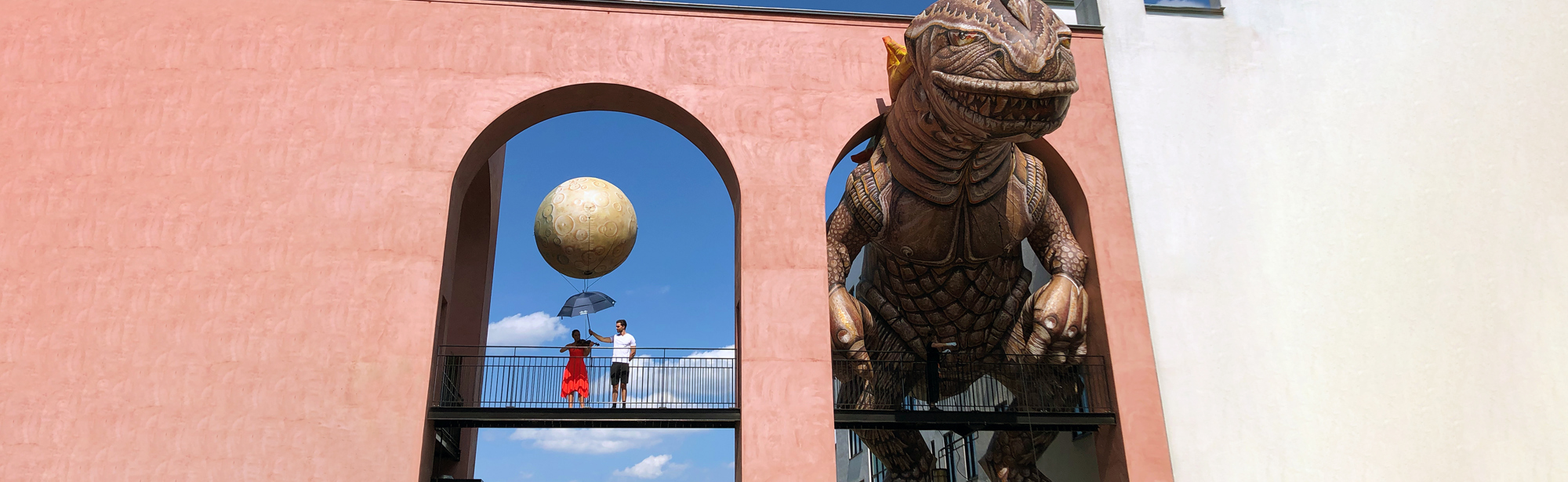 Grote muur van gebouw met daarin twee grote openingen waarin de ene boog 2 mensen die een grote ballon vasthouden en in de andere boog een enorme dino komt.