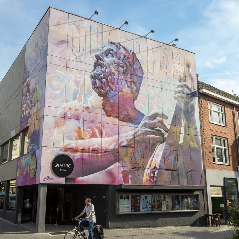 Een man op een fiets fietst langs de Quatro Cinema in Heerlen met een street art mural van een Romein