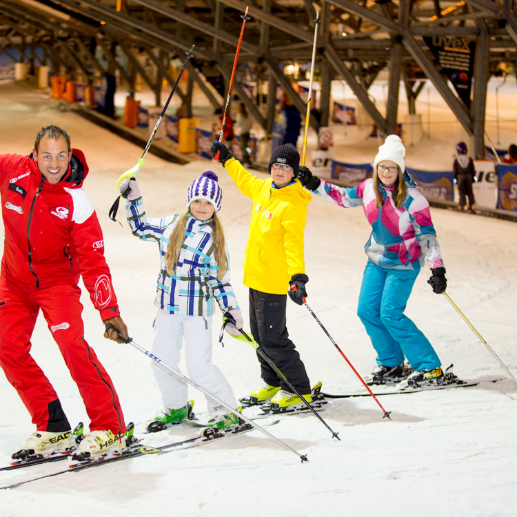 Een skileraar met drie kinderen skiën in een rijtje naar beneden met een skistok de lucht in 