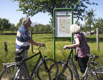 Twee ouderen mensen met fiets in hun hand kijken naar een infopaneel met fietsknooppunten