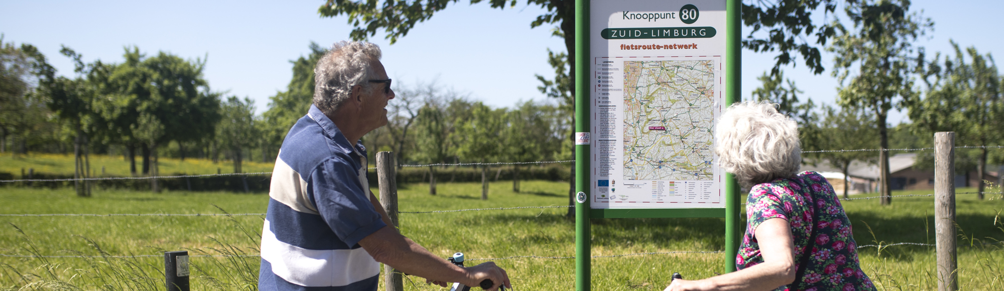 Twee ouderen mensen met fiets in hun hand kijken naar een infopaneel met fietsknooppunten