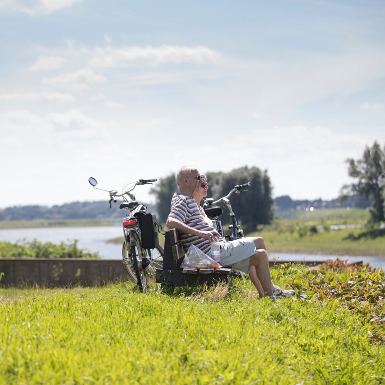 2 oudere mensen onderbreken hun fietstocht op een bankje op een zonnige dag in Rivierpark Maasvallei