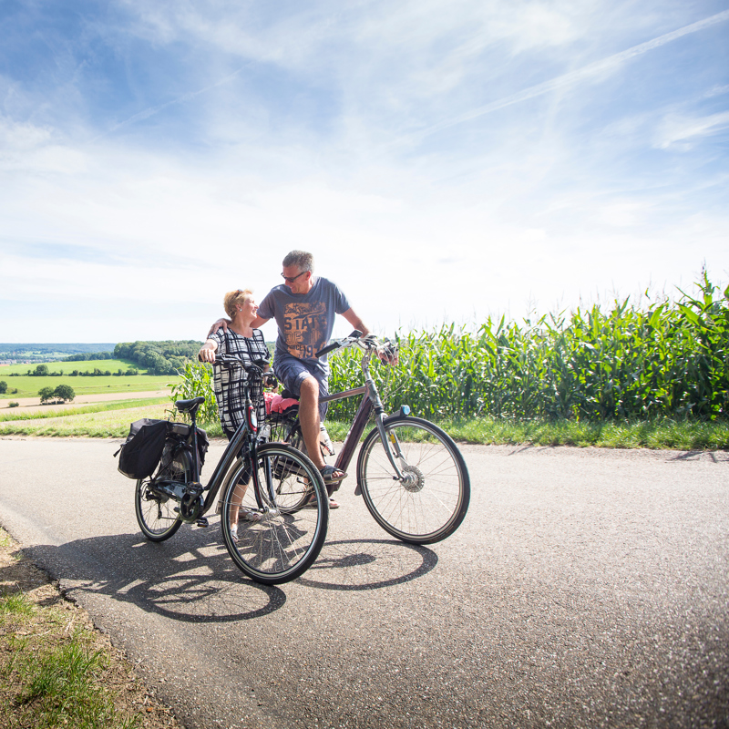 Een koppel op de fiets kijkt elkaar boven op de berg verliefd aan met achter zich een panorama over de heuvels