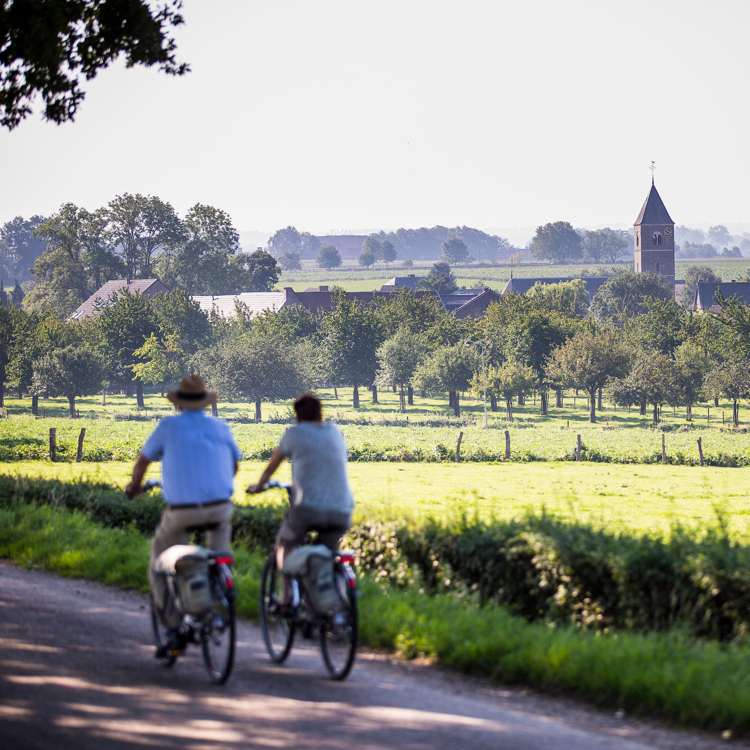 Koppel fietst langs weiland met uitzicht op de kerk en het dorpje Mesch