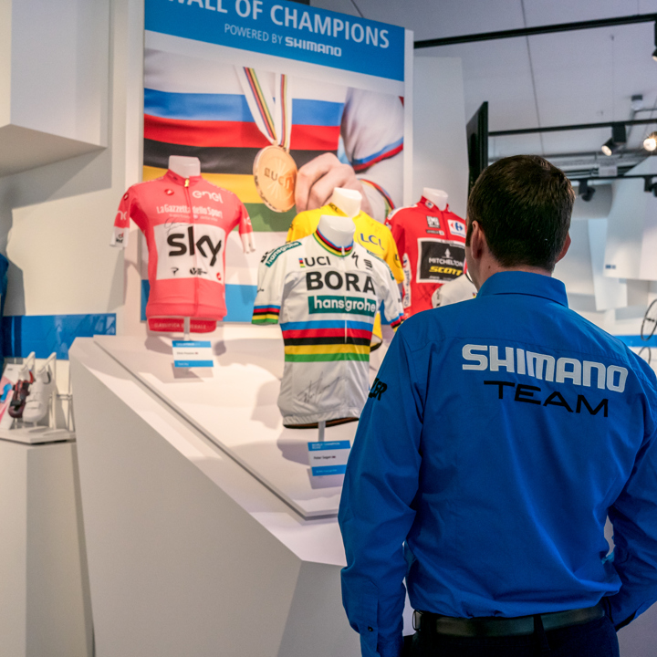 Man met Shimano vest kijkend naar borstbeelden met wielershirts van kampioenen in Shimano Experience Center