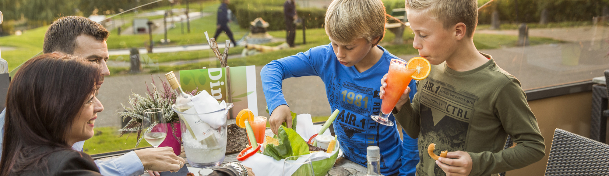 Ouders met twee kinderen drinken zomerse drankjes op het terras met uitzicht op de speeltuin