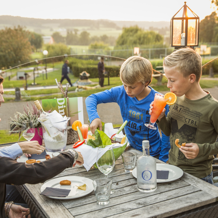 Ouders met twee kinderen drinken zomerse drankjes op het terras met uitzicht op de speeltuin