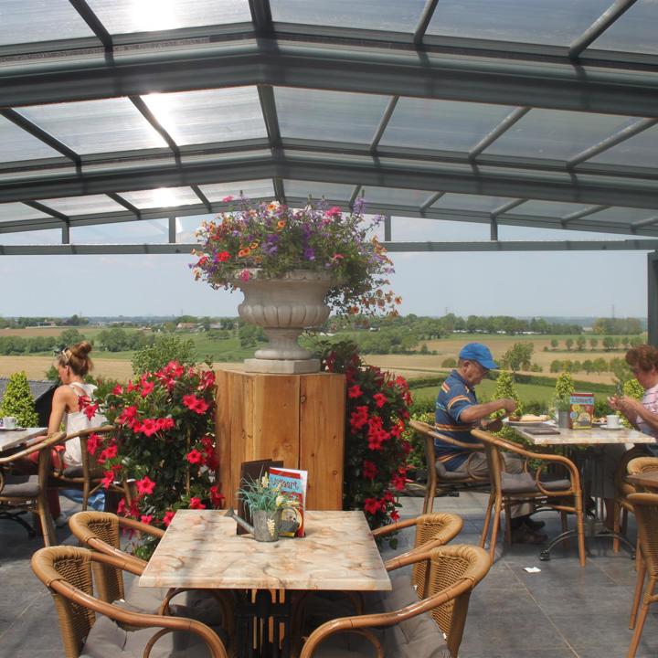 Een overdekt terras met open glazen pui waar twee koppels zitten te lunchen en uitkijken op een panorama