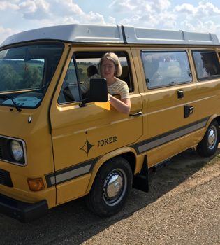 Vrouw zit achter het stuur van een geel camperbusje. 