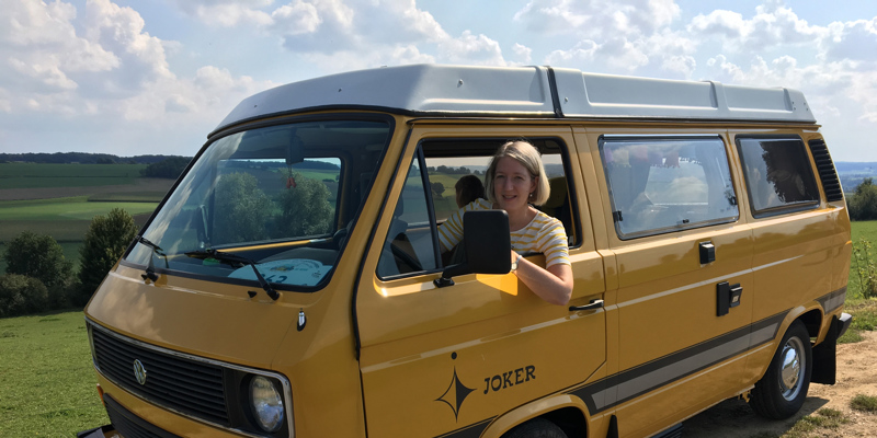 Vrouw zit achter het stuur van een geel camperbusje. 