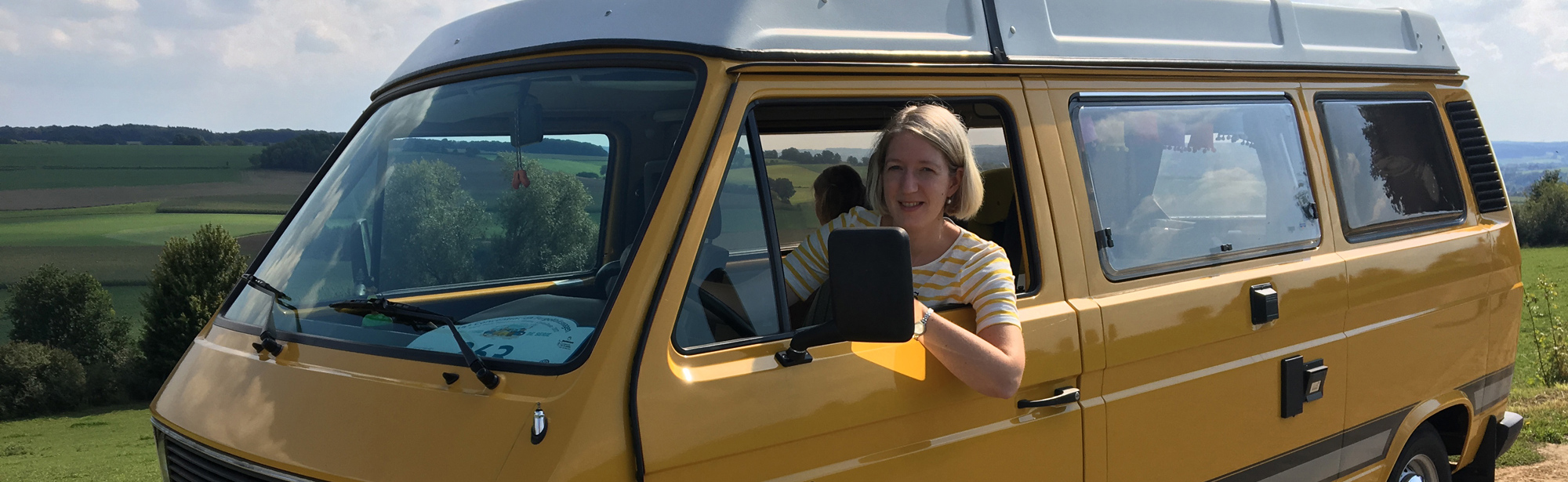 Vrouw zit achter het stuur van een geel camperbusje. 