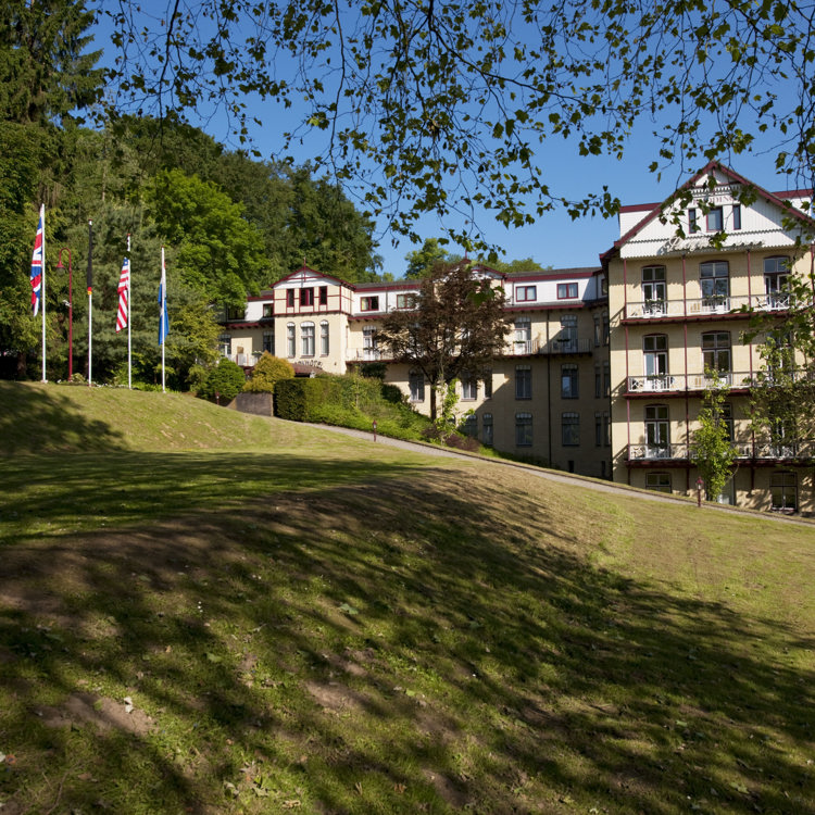 Groot hotelgebouw aan de rand van het bos op een heuvel met vlaggen van diverse landen. 