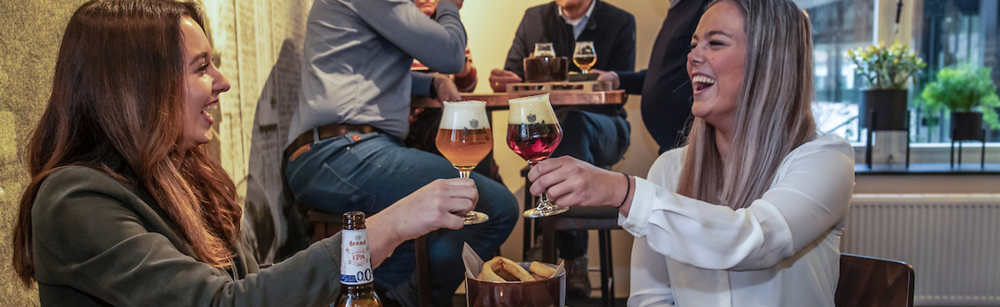 Twee vrouwen proosten al lachend met hun biertjes bij het Brand Proeflokaal met op de achtergrond andere gasten