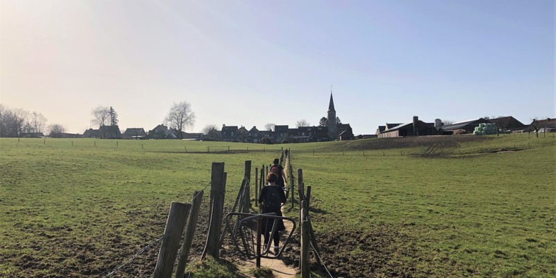 Twee vrouwen lopen over een wandelpad met draaihekjes door een weiland met op de achtergrond een dorpje en kerkje