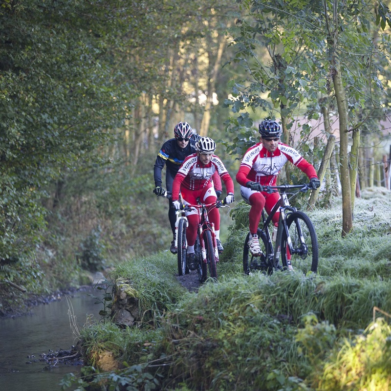 Een groep mountainbikers met rode en zwarte outfits fietsen over een bospad langs een beekje