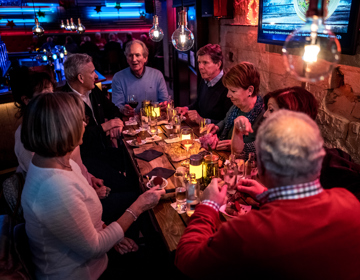 8 mensen zitten aan een lange houten tafel met hapjes en drankjes