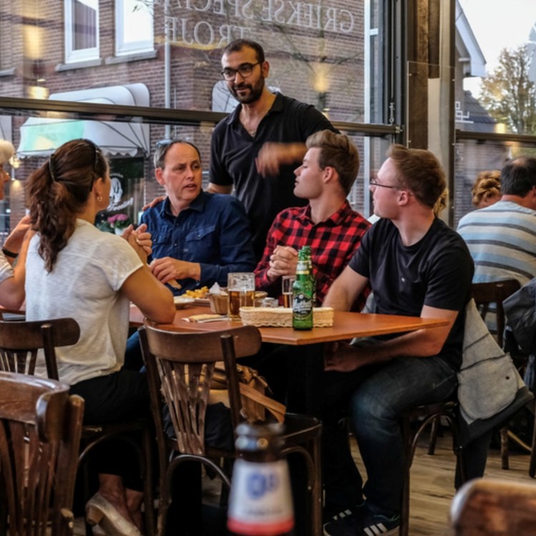 Een ober praat met een vijftal gasten aan een tafeltje aan het raam