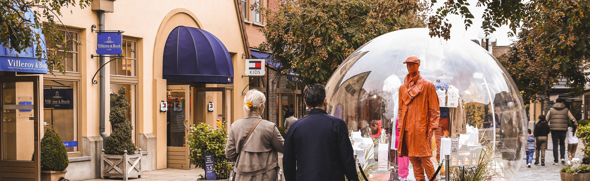 Twee mensen en een kindje lopen door de Maasmechelen Village Winkelstraat met een kunstwerk in het midden van de straat