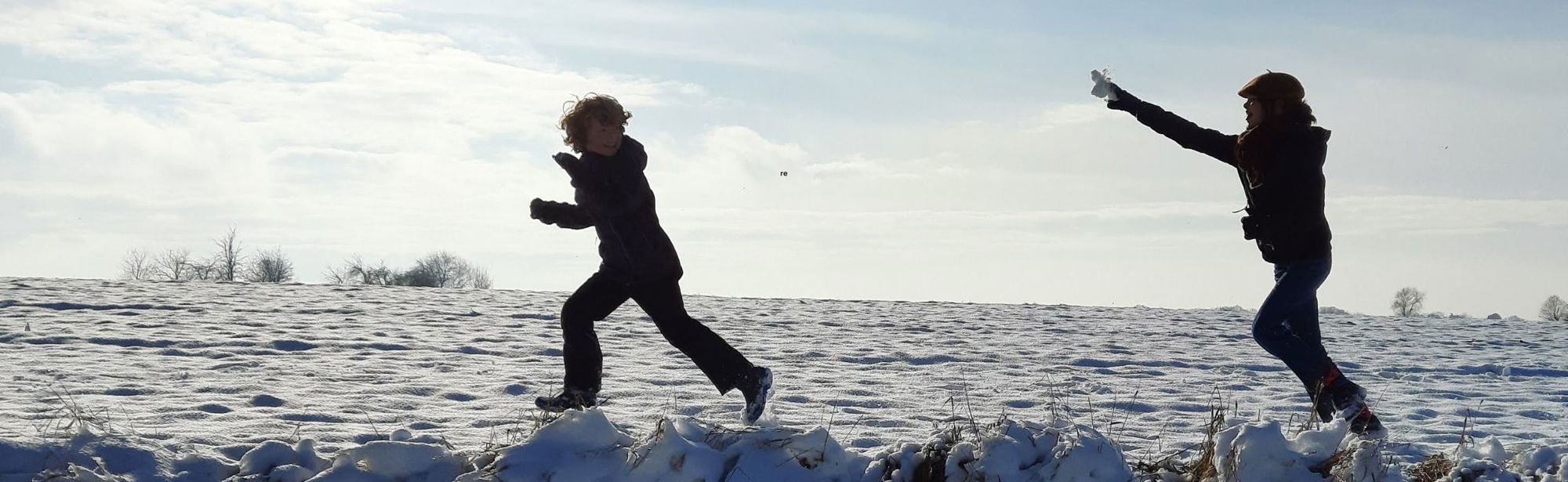 Twee kinderen rennen achter elkaar aan en gooien sneeuwballen in een besneeuwd landschap. 